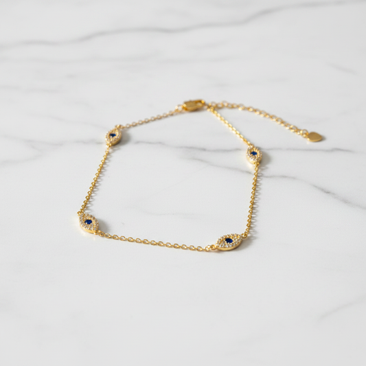 Evil eye gold anklet with pavé crystal charms on white marble surface, clean light background