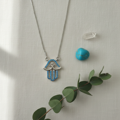 Boho flat lay of silver turquoise hamsa necklace on white linen with turquoise stone and eucalyptus