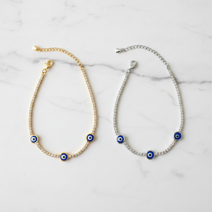 Gold and silver evil eye tennis bracelets side by side on white marble showcase