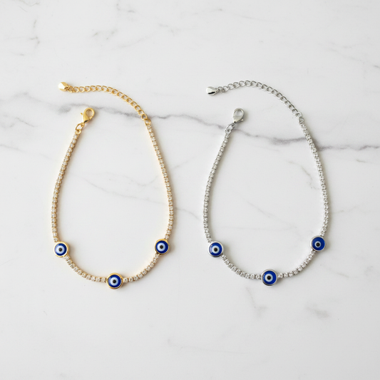 Gold and silver evil eye tennis bracelets side by side on white marble showcase